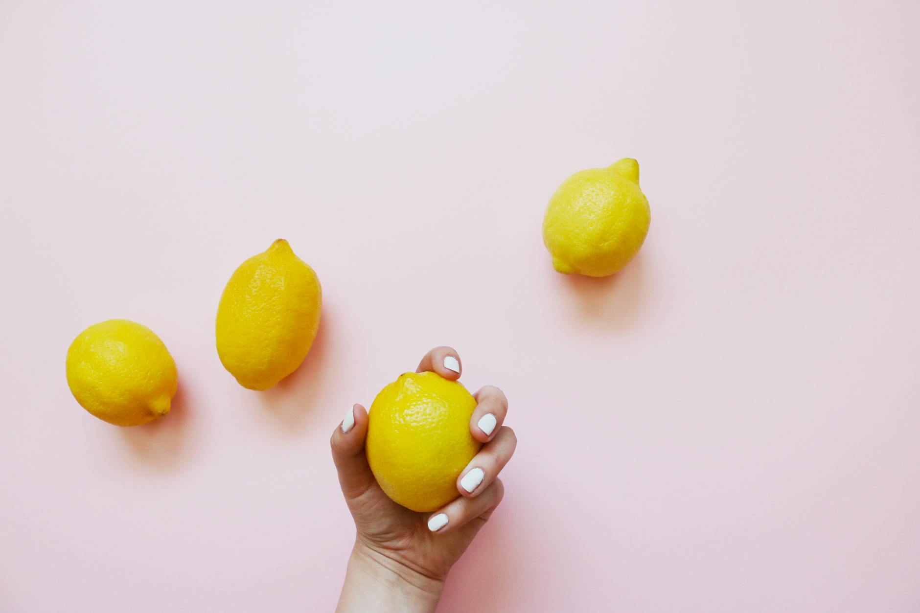 A hand holding a fresh lemon on a soft pink background surrounded by additional lemons