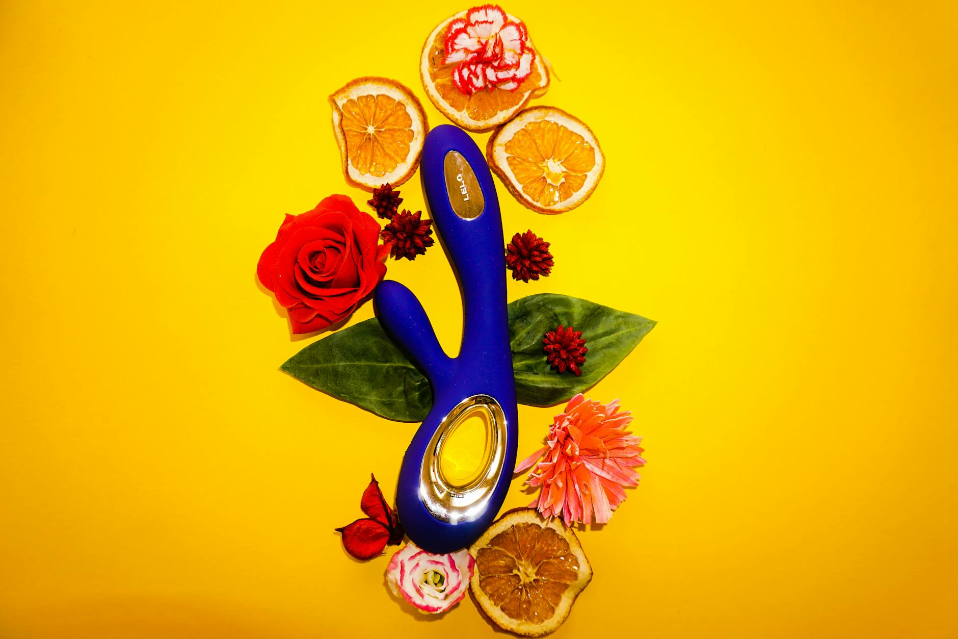 A blue vibrator surrounded by flowers and citrus on a bright yellow background, symbolizing fresh approaches to intimate reconnection.